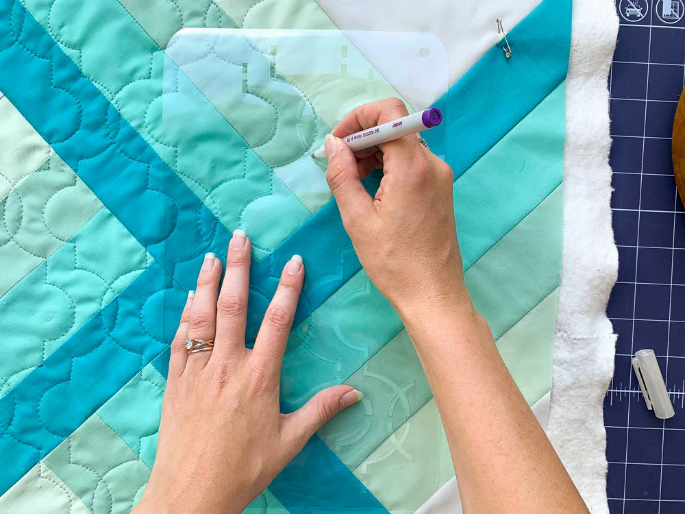 5 Minute Guide to Quilt Marking Tools: Hands trace the lines of a quilt stencil using an air and water-soluble pen on a blue Adventureland quilt. #quilting #sewingdiy suzyquilts.com