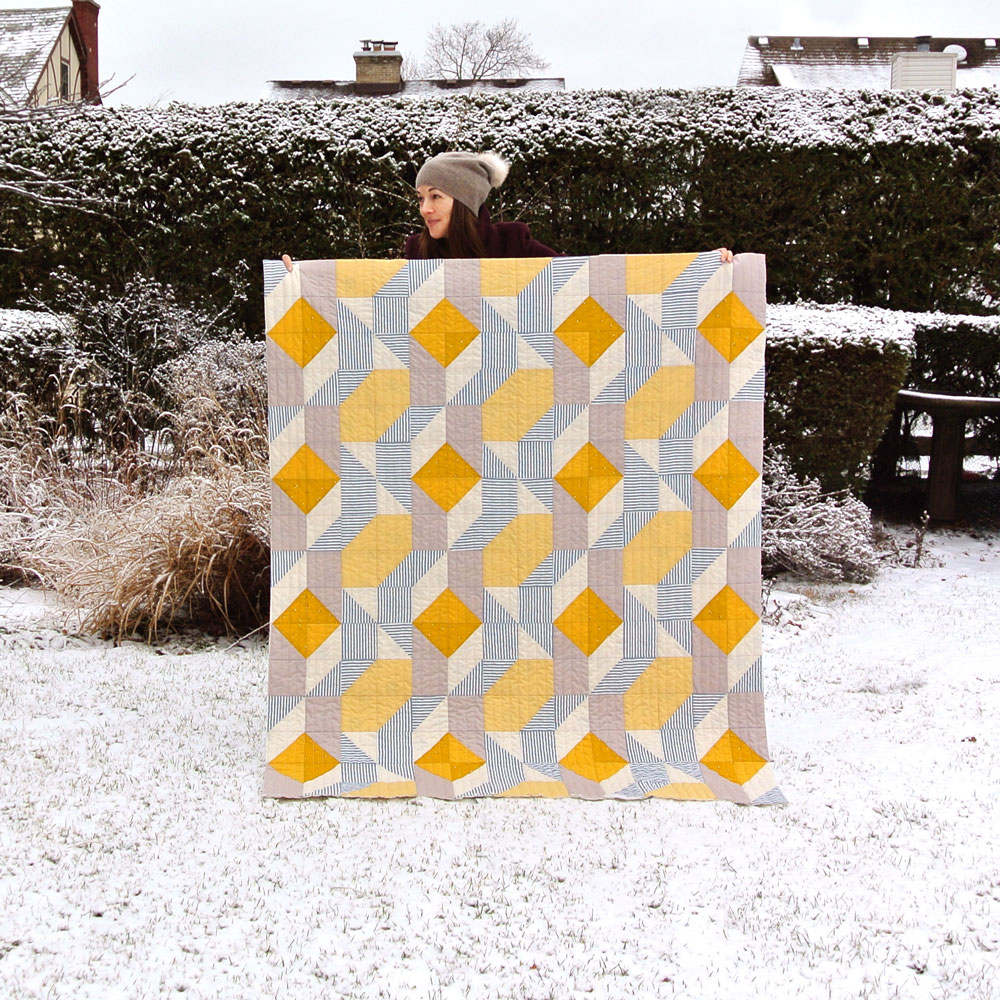 Easy Half Square Triangles Tutorial: A yellow and grey Rocksteady quilt held outside with a snowy background. suzyquilts.com #quilting #sewingdiy