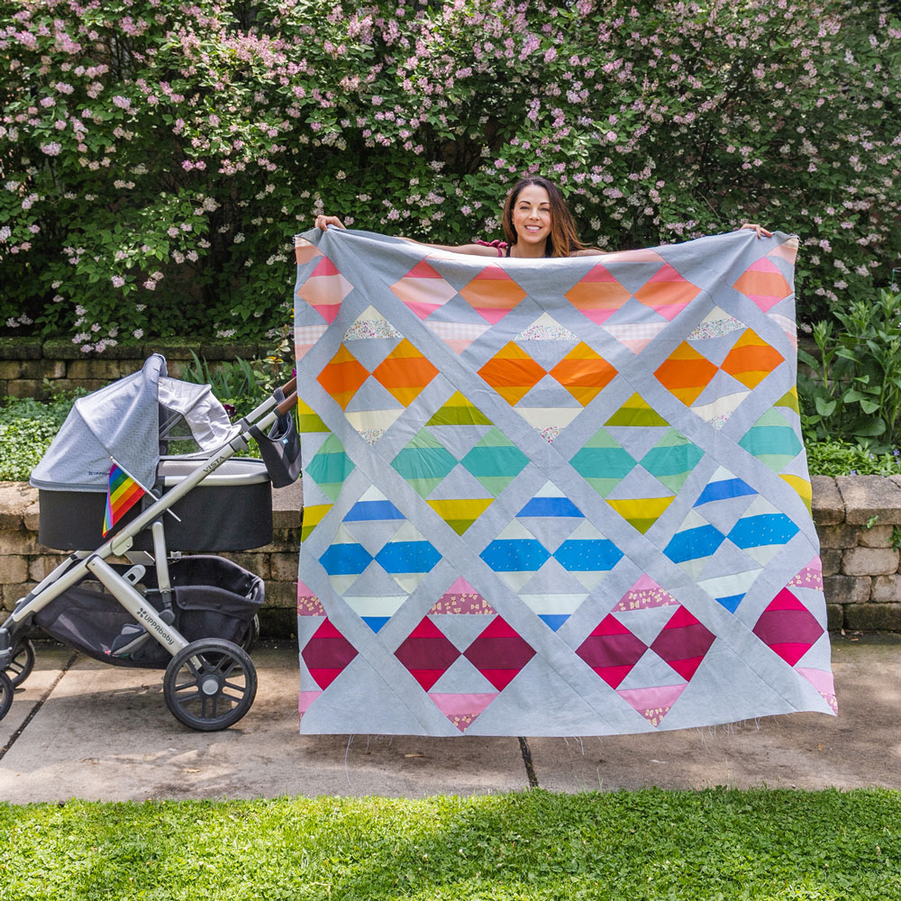 Easy Half Square Triangles Tutorial: A rainbow colored New Horizons quilt top with a grey background is held outside next to a stroller. suzyquilts.com #quilting #sewingdiy
