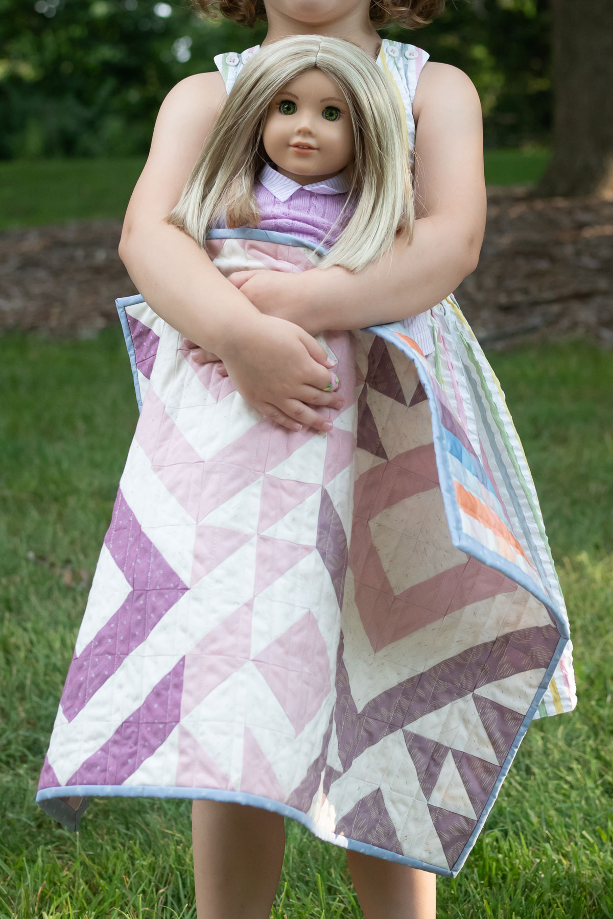 A little girl holds a doll that is wrapped in a pink and purple Triangle Jitters doll quilt. #Quilting #SewingDIY suzyquilts.com A little girl holds a doll that is wrapped in a pink and purple Triangle Jitters doll quilt. #Quilting #SewingDIY suzyquilts.com